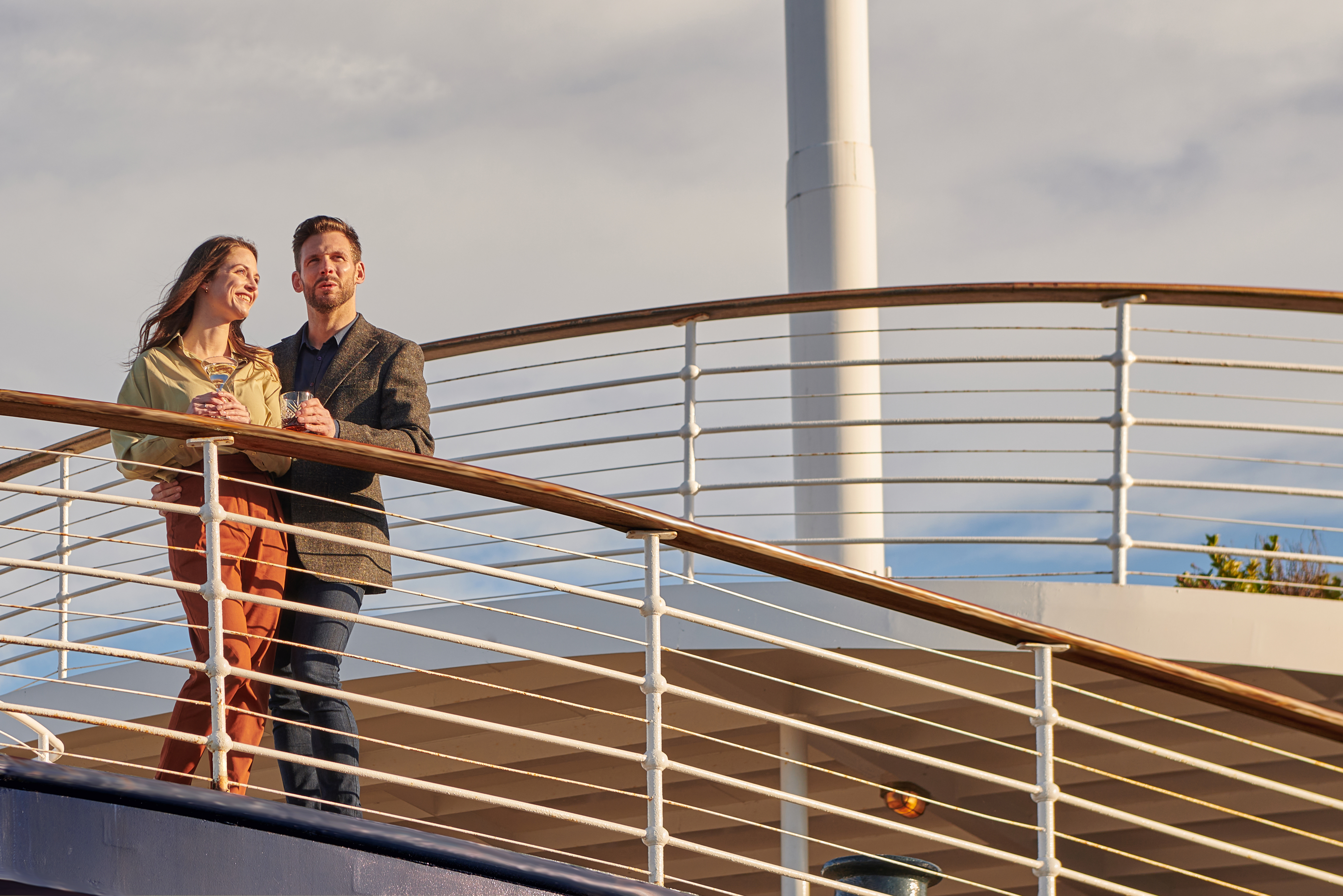 A man and woman stand, each with a drink in hand, looking out from the deck aboard Fingal