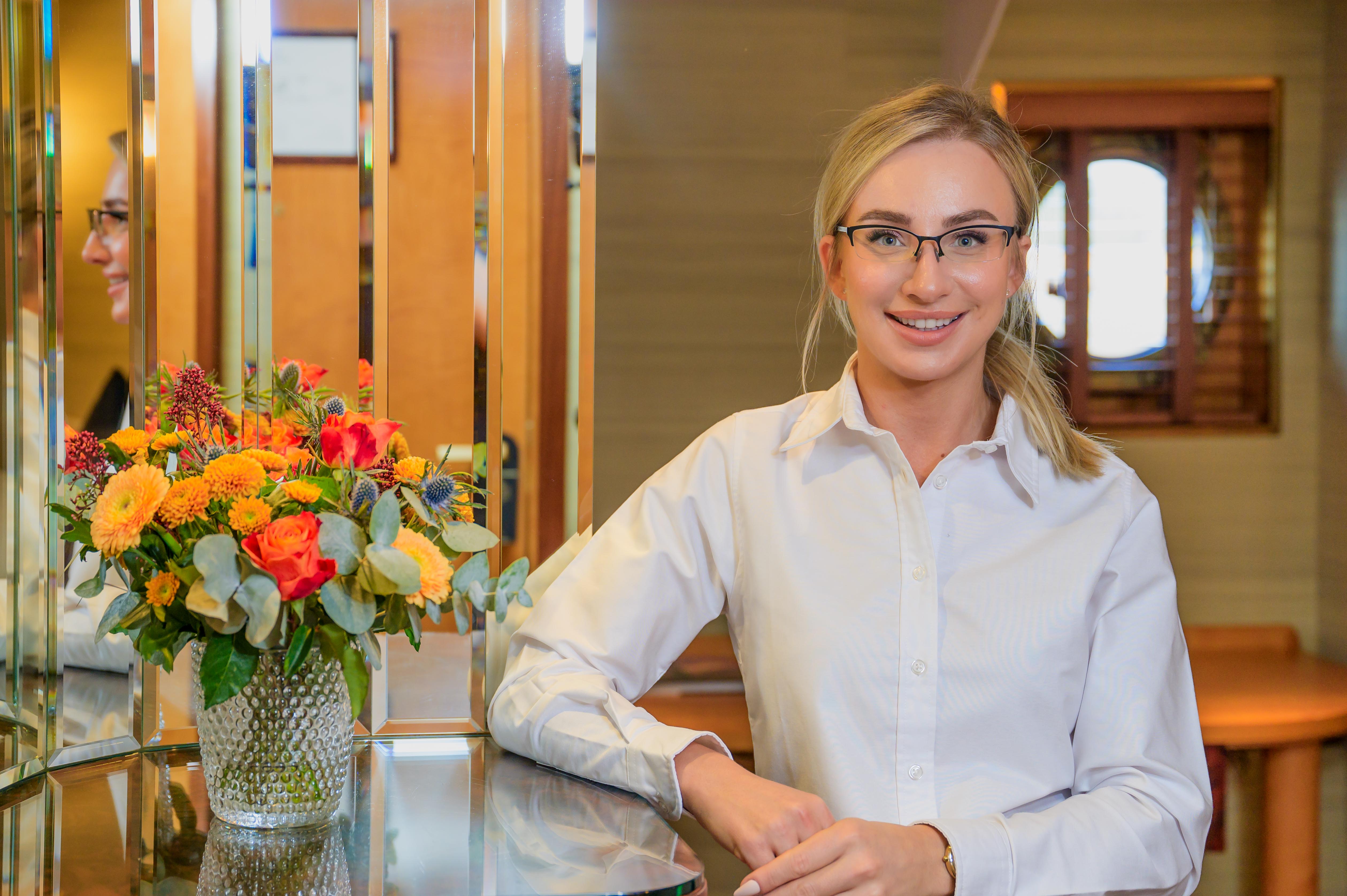 A woman wearing a white shirt is standing smiling next to a mirror in a cabin at Fingal Hotel. 