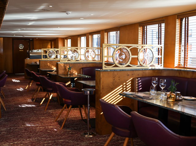 Interior shot of The Lighthouse restaurant aboard Fingal, an iconic floating hotel in Edinburgh, showcasing elegant tables set for fine dining with warm wood interiors, plush seating, and natural light streaming through the windows.
